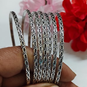 May include: A set of silver bangles with an intricate oval and cross pattern. The bangles are displayed against a soft background with pink and red flowers. The design is repeated along the length of each bangle.