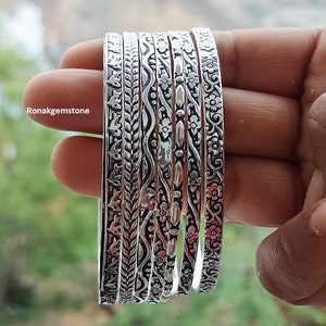 May include: A collection of silver bangles with intricate floral and leaf patterns. The bangles are stacked together, showcasing detailed designs and textures. The image includes the text "Ronakgemstone".