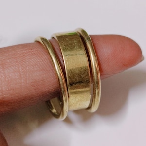 Peut inclure: Une bague dorée avec une bande centrale rectangulaire épaisse et deux bandes arrondies plus fines de chaque côté. La bague est portée sur un doigt, mettant en valeur son design simple mais élégant. La surface polie de la bague reflète la lumière.