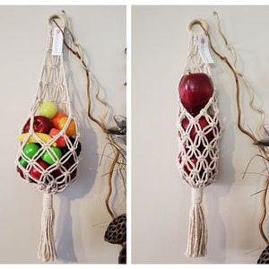 May include: Two macrame hanging fruit baskets. The basket on the left is filled with a variety of colourful fruit. The basket on the right is filled with red apples.