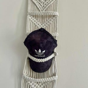 May include: A cream macrame wall hanging with a black Adidas baseball cap. The macrame features a geometric design and pockets. The cap displays the Adidas logo in white.