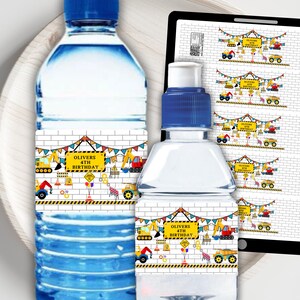 Op de afbeelding: Twee doorzichtige plastic waterflessen met aangepaste labels met een bouwthema, inclusief gele graafmachines, verkeerskegels en kleurrijke vlaggetjes. De labels tonen de tekst "Olivers 4th Birthday" op een geel bord tegen een witte bakstenen achtergrond.