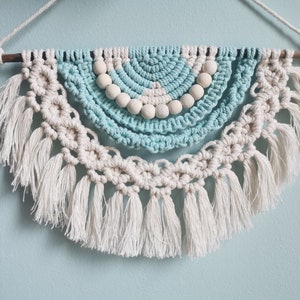 Könnte beinhalten: Ein Makramee-Wandbehang mit einem Halbkreis-Design. Das Stück hat ein hellblaues und cremefarbenes Farbschema mit Holzperlen und Fransen. Es wird an einem natürlichen Holzdübel und einer weißen Kordel aufgehängt.