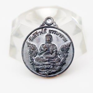 May include: A round, bronze-colored pendant with a depiction of a seated Buddha. The pendant has Thai text around the edge that reads "พระพุทธชินราช"