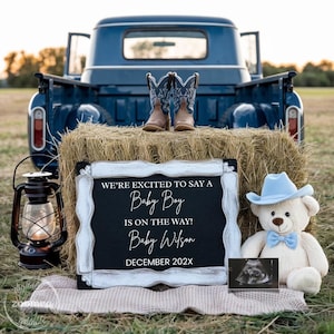 May include: A blue vintage pickup truck bed holds a bale of hay with cowboy boots. A chalkboard sign reads "We're excited to say a Baby Boy is on the way! Baby Wilson December 202X." A teddy bear with a blue hat and bow tie sits nearby.