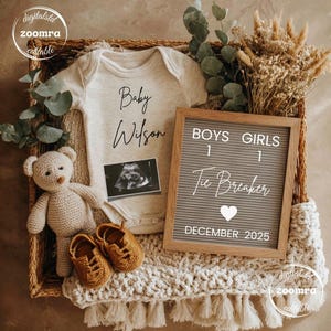 May include: A neutral-toned baby announcement arrangement. It includes a onesie with "Baby Wilson" printed on it, a sonogram, a letter board with "Boys 1 Girls 1 Tie Breaker December 2025", a teddy bear, and baby shoes.
