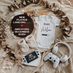 Puede incluir: Anuncio de bebé con un body blanco con la frase "BABY Wilson", un letrero de madera que dice "NEW PLAYER LOADING... ARRIVING DECEMBER 202X", un dispositivo de juego portátil, un controlador, auriculares y botines. El tema es un bebé gamer.