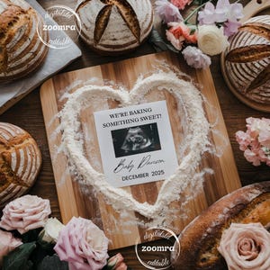 May include: A wooden cutting board with flour shaped into a heart, surrounding a card that reads "WE'RE BAKING SOMETHING SWEET!" with a sonogram image and "Baby Davison DECEMBER 2025". Several loaves of bread and flowers are arranged around the board.