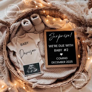 May include: A beige baby onesie with the name "Davison" and a sonogram image are displayed in a woven basket. A black letter board reads "Surprise! We're due with Baby #2 Coming December 2025." Beige baby booties are also in the basket.