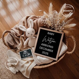 May include: A woven basket filled with a beige blanket, a white stuffed bunny, and a black letter board. The letter board reads "BABY Watson ARRIVING DECEMBER 2025". A sonogram image and dried flowers are also in the basket.