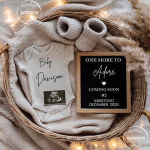 Peut inclure: Un body de bébé beige avec le texte "Baby Davison" et une image d'échographie, accompagné de chaussons tricotés et d'un tableau de lettres annonçant une arrivée en décembre 2025. Le tout est disposé dans un panier tressé.