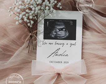 May include: A white card with a black and white ultrasound image of a baby. The text on the card reads "We are having a girl Amelia DECEMBER 2025". The card is decorated with a pink bow.