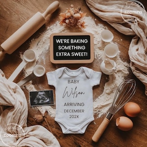 May include: A charming announcement featuring baking-themed elements. A white onesie with "Baby Wilson" and arrival details is surrounded by a rolling pin, whisk, eggs, and flour. A chalkboard reads "We're Baking Something Extra Sweet!"