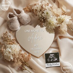Puede incluir: Un espejo en forma de corazón con el texto "ONE MORE TO Adore #2 DECEMBER 2025" se encuentra entre flores y patucos. Una foto de ultrasonido dice "Baby Wilson". La escena está ambientada sobre una tela color crema.
