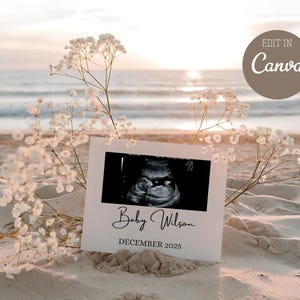 Op de afbeelding: Een echografie van een baby, weergegeven op een witte kaart met de tekst "Baby Wilson" en "DECEMBER 2025". De kaart staat op een zandstrand met witte bloemen en een zonsondergang op de achtergrond. De woorden "EDIT IN Canva" staan in een bruine cirkel.