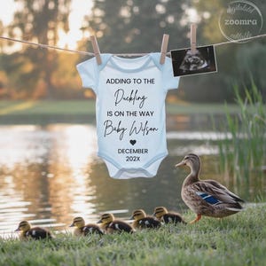 Puede incluir: Un body de bebé azul claro con el texto "Adding to the Duckling is on the way Baby Wilson December 202X" colgado de un tendedero. Una imagen de ultrasonido está sujeta a la línea. Una madre pato y patitos están en primer plano.