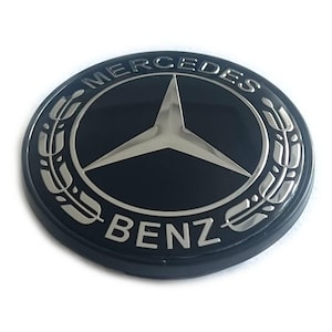 Puede incluir: Un emblema circular negro y plateado con el logotipo de Mercedes-Benz. El diseño incluye una estrella central de tres puntas rodeada por un anillo con el texto MERCEDES en la parte superior y BENZ en la inferior, flanqueado por hojas de laurel.
