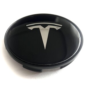 Peut inclure: Un cache-moyeu de roue circulaire avec une finition noire brillante et un logo Tesla argenté centré. Le capuchon présente une surface supérieure lisse et arrondie ainsi qu'une base en plastique noir mat avec des clips de fixation visibles.