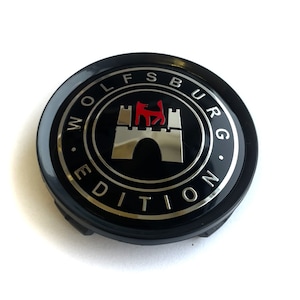 Könnte beinhalten: Rundes schwarzes Autoemblem mit silberner Einfassung und den Worten "Wolfsburg Edition" um ein silbernes Schloss mit einem roten Wolf darauf.