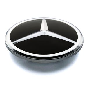 May include: A silver and black Mercedes-Benz emblem. The emblem features a three-pointed star in silver, set against a black background, within a silver circular border. The emblem is slightly raised.