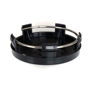 May include: A circular black plastic wheel center cap featuring a smooth, glossy finish. The component includes four evenly spaced mounting clips along the perimeter and a thin, circular silver metal tension ring secured around the clips.