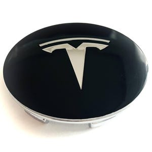 Peut inclure: Un cache-moyeu de roue circulaire avec une finition noire brillante et un emblème Tesla argenté proéminent centré sur la surface. Le cache possède une base en argent métallique avec des clips de fixation visibles sur le bord inférieur.