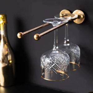 Può includere: Un porta bicchieri da vino in metallo dorato montato su una parete nera con due bicchieri da vino appesi.