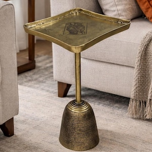 May include: A gold-colored side table with a square top and a decorative base. The table has a long, slender stem and a patterned, bell-shaped base. The top has a design in the center. The table is placed on a patterned rug.
