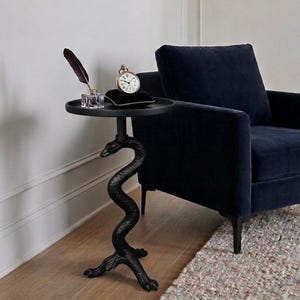 May include: A black side table featuring a circular top supported by a sculptural base shaped like a coiled snake. On the table sits a clear glass inkwell with a dark feather quill and a silver pocket watch resting on a small black velvet cushion.