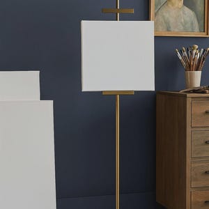 May include: A gold-colored metal artist's easel with a white canvas. The easel has a vertical pole and a rectangular base. A painting and a wooden cabinet are in the background.