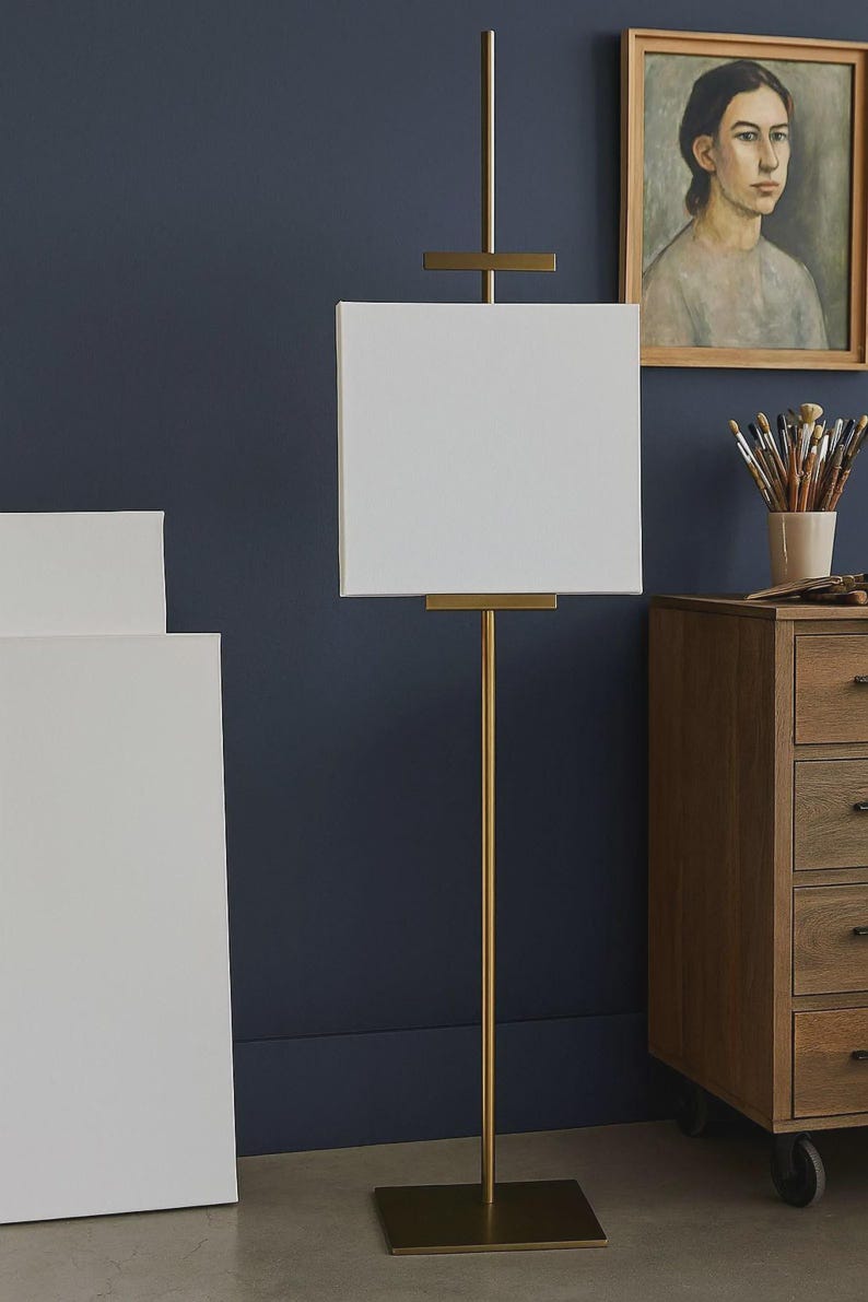 May include: A gold-colored metal artist's easel with a white canvas. The easel has a vertical pole and a rectangular base. A painting and a wooden cabinet are in the background.