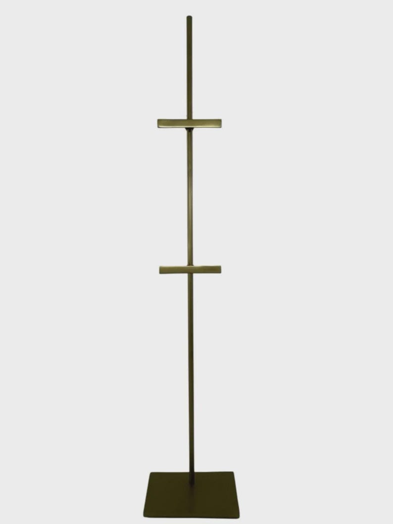 May include: A tall, bronze-colored display stand with a square base and a vertical pole. Two horizontal bars extend from the pole, creating a T-shape. The stand is designed to hold items for display, such as artwork or signage.