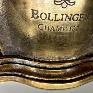 May include: A gold-colored metal ice bucket with a decorative design. The bucket has the text "BOLLINGER CHAMP 1872" engraved on the front.