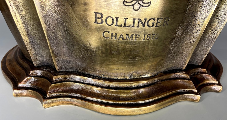 May include: A gold-colored metal ice bucket with a decorative design. The bucket has the text "BOLLINGER CHAMP 1872" engraved on the front.