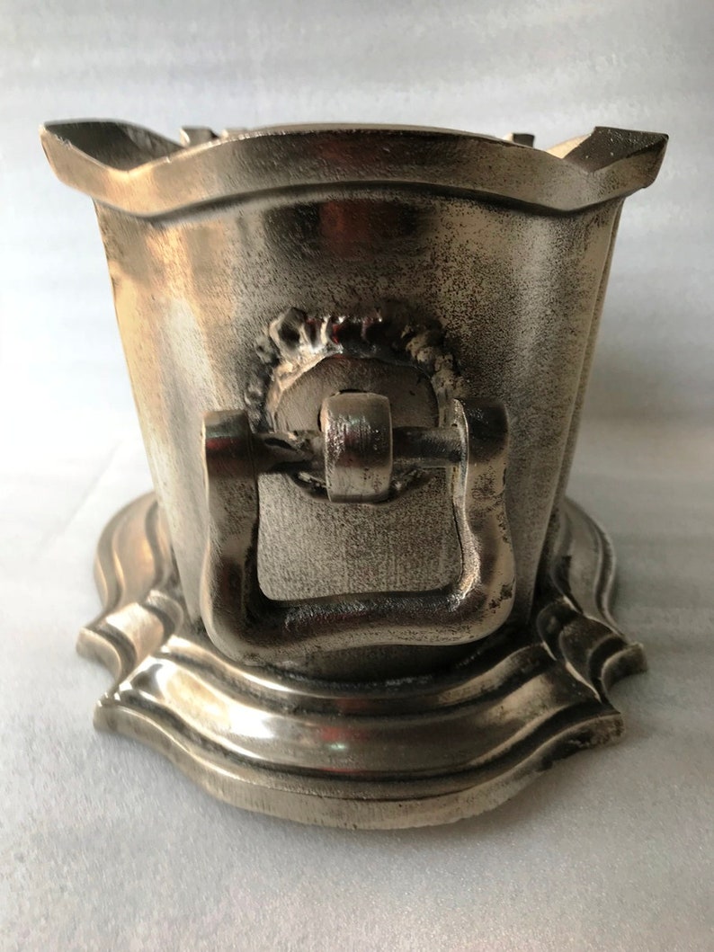 May include: A silver metal ice bucket with a decorative handle and a scalloped base.