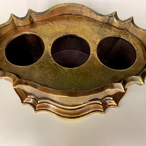 May include: A gold-toned metal decorative stand with a round top and three circular openings. The stand has a scalloped edge and a decorative base.