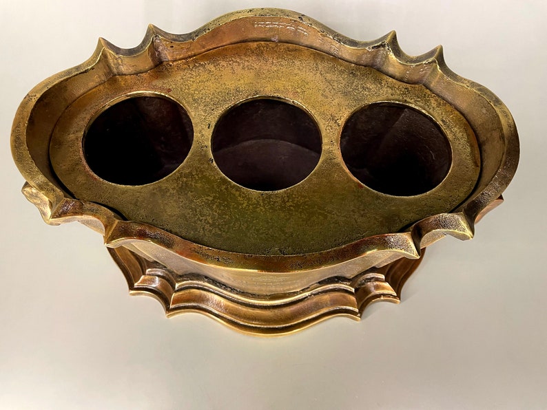 May include: A gold-toned metal decorative stand with a round top and three circular openings. The stand has a scalloped edge and a decorative base.