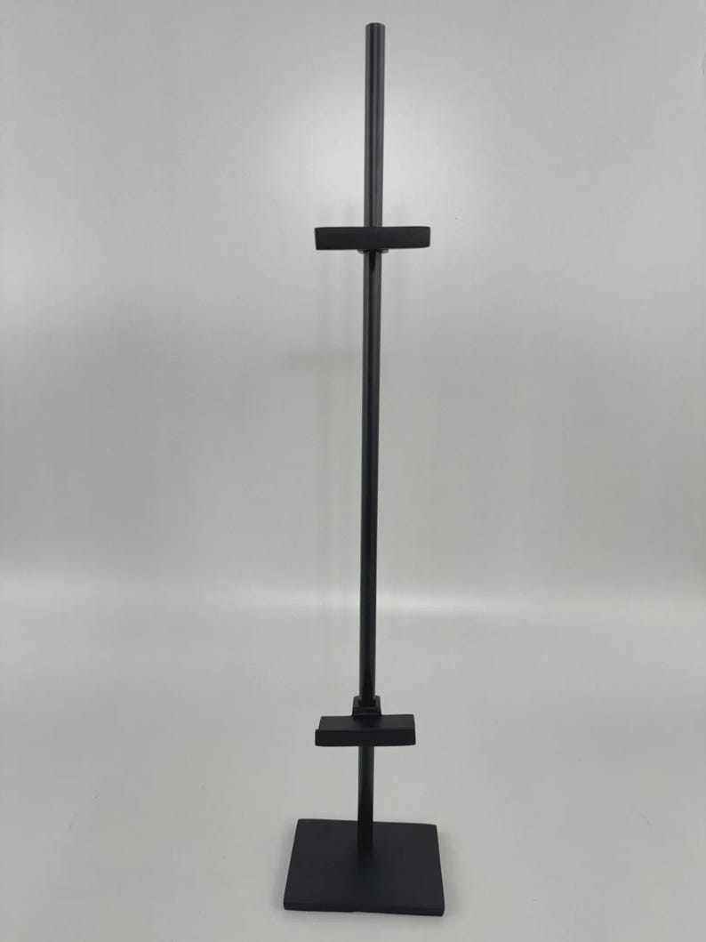 May include: A black metal display stand with a square base, a vertical pole, and two horizontal crossbars. The stand is designed to hold items for display. The stand is made of metal and has a matte finish.