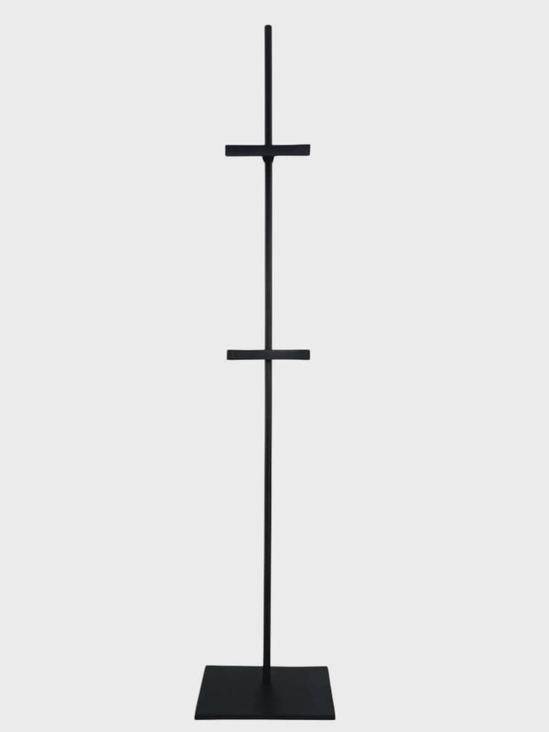 May include: A tall, black metal easel with a square base and two horizontal bars. The easel is designed to hold a canvas or artwork, and it stands against a plain white background.