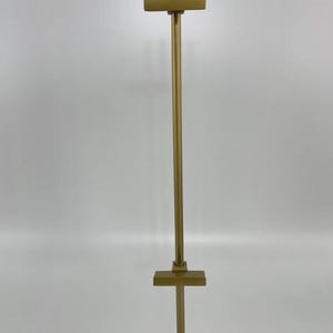 May include: A tall, gold-colored display stand with a square base and a vertical pole. The pole has horizontal crossbars at the top and middle, creating a T-shape. The stand is designed to hold items for display.