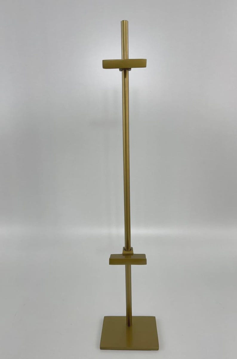 May include: A tall, gold-colored display stand with a square base and a vertical pole. The pole has horizontal crossbars at the top and middle, creating a T-shape. The stand is designed to hold items for display.