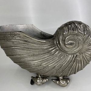 Op de afbeelding: Een zilveren metalen schelpvormige plantenbak met een getextureerd oppervlak en drie poten. De plantenbak is gevormd als een schelp en heeft een spiraalvormig ontwerp.
