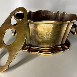 May include: A gold metal champagne bucket with a removable lid. The lid has three holes for holding bottles. The bucket has a decorative design and the text "Bollinger Champagne" engraved on the front.