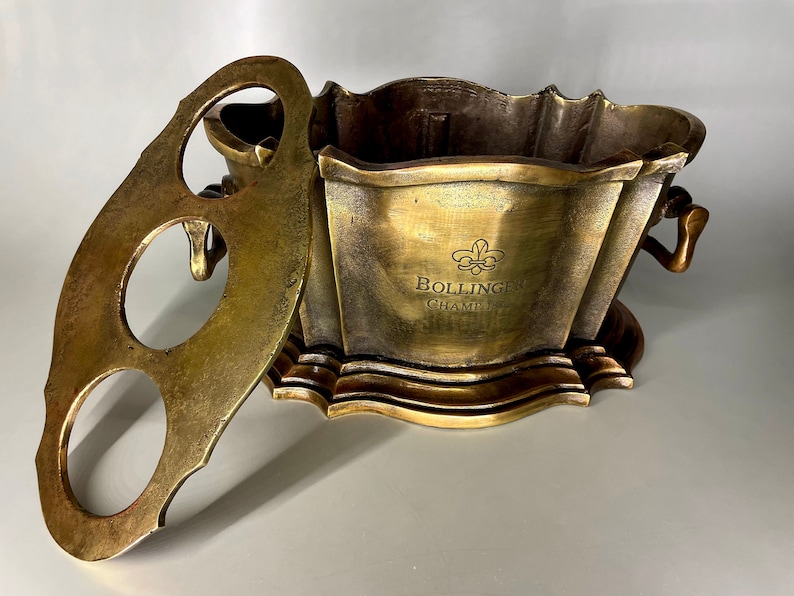 May include: A gold metal champagne bucket with a removable lid. The lid has three holes for holding bottles. The bucket has a decorative design and the text "Bollinger Champagne" engraved on the front.