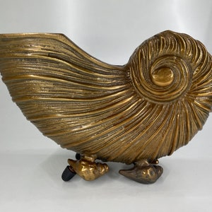 Op de afbeelding: Een gouden metalen schelpvormige plantenpot met een gedetailleerd, getextureerd oppervlak en drie poten.