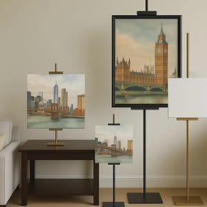 Könnte beinhalten: Verschiedene Größen von schwarzen und goldenen Staffeleien mit Stadtlandschaftsbildern. Ein Gemälde zeigt die Brooklyn Bridge und die Skyline von New York. Ein anderes zeigt Big Ben und das Parlament.
