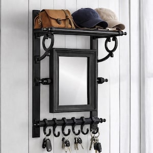 May include: Black metal wall-mounted organizer with a shelf holding a brown satchel and two caps. It features a rectangular mirror, decorative scrollwork, and hooks with keys hanging from them. The organizer is mounted on a white paneled wall.