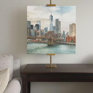 May include: Watercolor painting of the New York City skyline, including the Brooklyn Bridge and One World Trade Center. The artwork is displayed on a gold-colored easel on a dark brown table. The painting features shades of blue, brown, and gray.