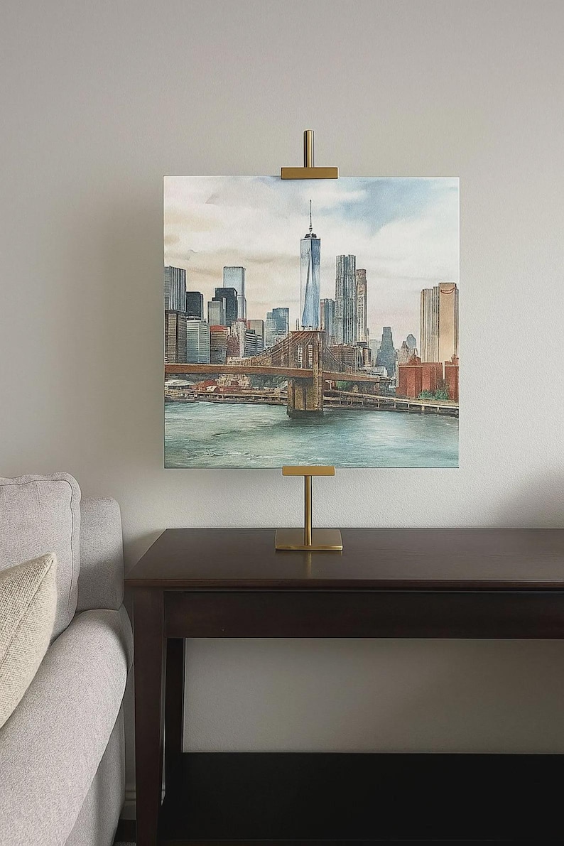 May include: Watercolor painting of the New York City skyline, including the Brooklyn Bridge and One World Trade Center. The artwork is displayed on a gold-colored easel on a dark brown table. The painting features shades of blue, brown, and gray.