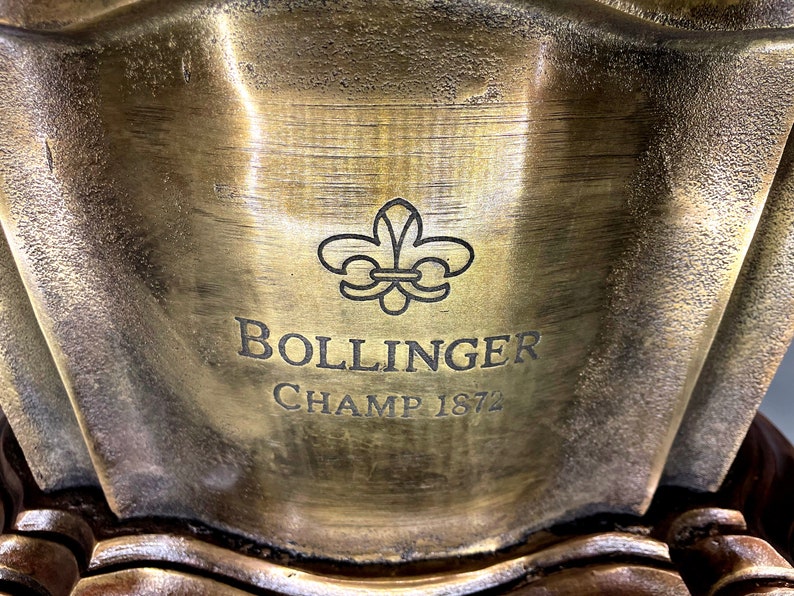 May include: A gold-colored metal plaque with a fleur-de-lis design and the text "Bollinger Champ 1872".